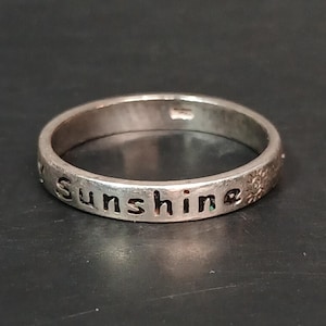 以下が含まれることがあります： 「sunshine」の文字が刻まれたシルバーリング。シンプルなバンドスタイルで、少し使い古された外観です。リングは暗い表面に置かれています。