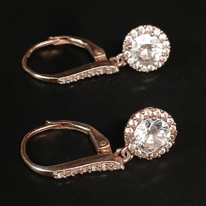 May include: A pair of rose gold leverback earrings. Each earring features a round, clear gemstone surrounded by smaller, clear gemstones. The leverback closure and the connecting bar are also adorned with small, clear gemstones. The earrings are displayed against a black background.