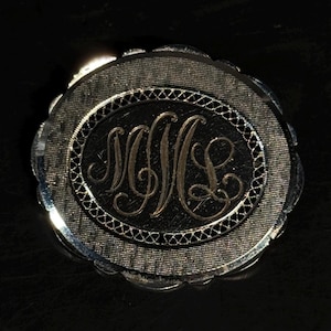 May include: A round, metallic brooch featuring a central oval panel with an engraved, cursive monogram of the letters SML. The piece has a textured, cross-hatched border surrounding the center and a scalloped, polished outer edge.