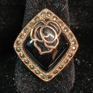 May include: A gold-toned ring with a black square center stone. A rose design is in the center of the black stone. The ring is set with small, green-toned stones around the black center stone.