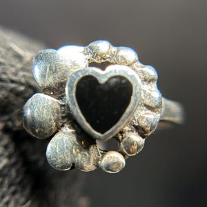 May include: A silver ring featuring a black heart-shaped stone at the center, surrounded by a decorative, textured border. The ring has a vintage or antique appearance, with a detailed design.