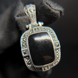 May include: A silver pendant with a rectangular black stone center. The pendant has a decorative border with small, sparkling accents. The bail is detailed with a textured design. The jewelry is displayed against a dark background.