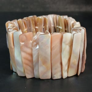 May include: A stretch bracelet made of rectangular, iridescent shell pieces in shades of peach, cream, and brown. The bracelet is designed to fit snugly around the wrist and has a natural, organic appearance. The shell pieces are strung together.