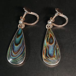 Puede incluir: Un par de pendientes de clip plateados con conchas de abulón en forma de lágrima. Las conchas muestran iridiscencias azules, verdes y marrones. Los pendientes tienen un mecanismo de rosca.