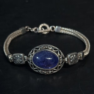 May include: Silver bracelet featuring a large, oval, deep blue gemstone at the center. The gemstone is set in an ornate, openwork silver frame. The bracelet has a woven chain design and a toggle clasp. The bracelet is displayed against a dark background.