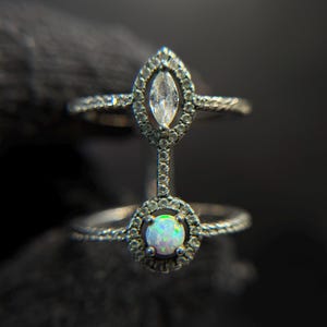 May include: Two-piece silver ring set. The top ring features a marquise-cut clear stone surrounded by smaller stones. The bottom ring has a round opal-like stone, also encircled by smaller stones. The rings are connected by a delicate bar.