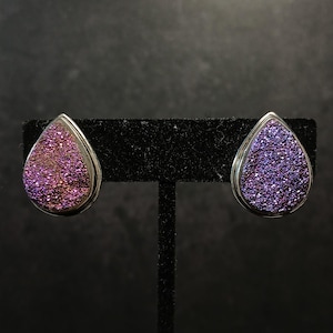 May include: Pair of teardrop-shaped earrings with a silver-colored border. The earrings have a sparkling, textured surface with shades of purple and pink. The earrings are displayed on a black stand against a dark background.