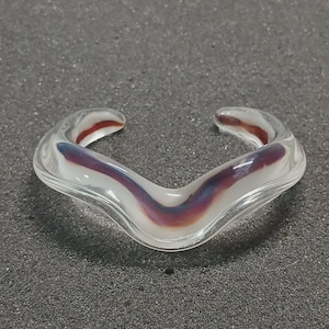 May include: A clear glass ring with a V-shaped design. The ring features a white base with streaks of purple and red. The open ends of the ring have red accents. The ring is on a dark gray surface.
