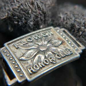 May include: Close-up of a silver-colored metal pin with the words "FLOWER HONOR CLUB" and a flower design. The pin is rectangular with a decorative border and is set against a dark, textured background.