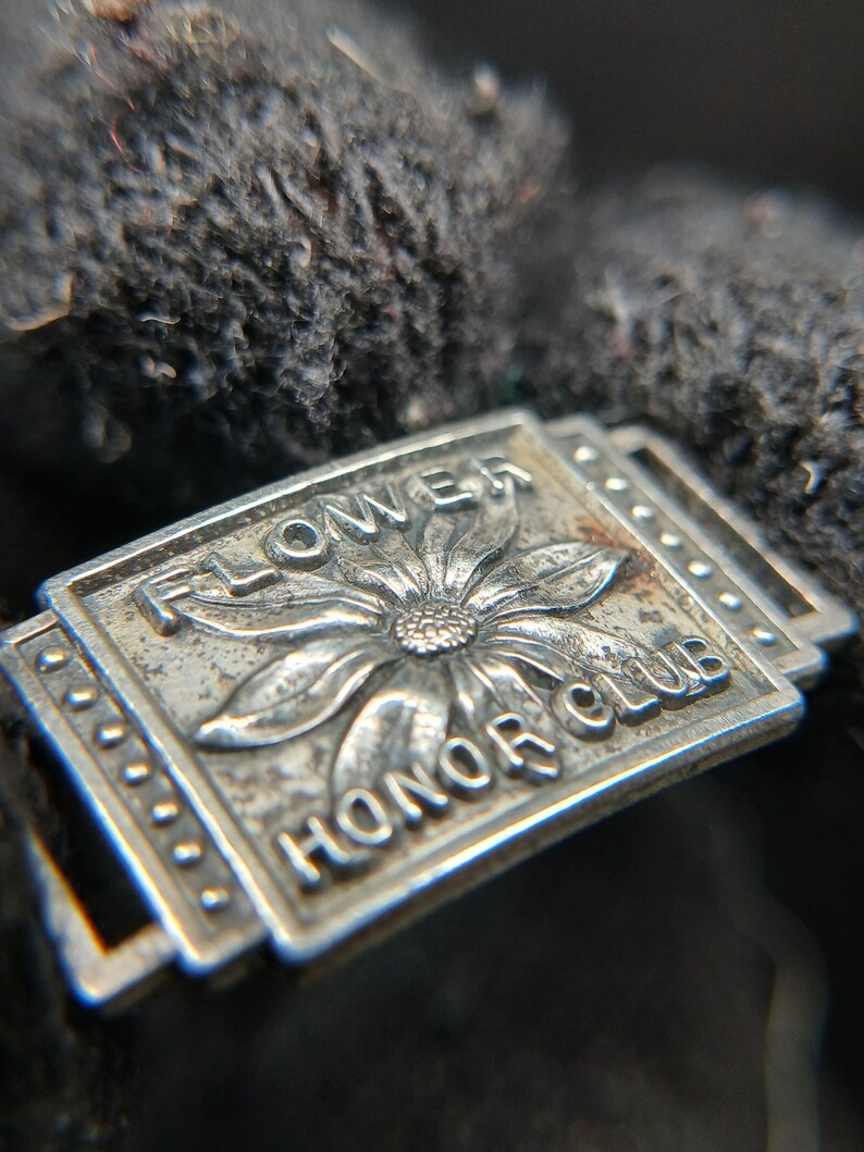 May include: Close-up of a silver-colored metal pin with the words "FLOWER HONOR CLUB" and a flower design. The pin is rectangular with a decorative border and is set against a dark, textured background.
