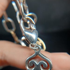 Puede incluir: Primer plano de un collar de plata con un cierre en forma de coraz&oacute;n y eslabones decorativos. El collar presenta un dise&ntilde;o detallado y ornamentado, con un enfoque en el cierre y los eslabones de conexi&oacute;n. El fondo es oscuro y borroso.