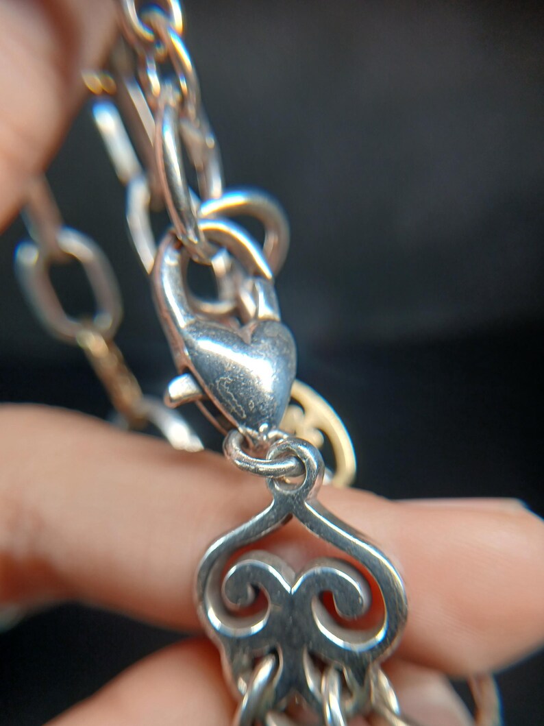 Puede incluir: Primer plano de un collar de plata con un cierre en forma de coraz&oacute;n y eslabones decorativos. El collar presenta un dise&ntilde;o detallado y ornamentado, con un enfoque en el cierre y los eslabones de conexi&oacute;n. El fondo es oscuro y borroso.