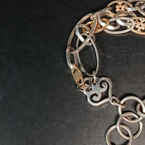Puede incluir: Una pulsera plateada y dorada con una variedad de estilos de eslabones. La pulsera presenta eslabones ovalados, circulares y decorativos, incluido un cierre en forma de coraz&oacute;n. El metal tiene un acabado pulido.