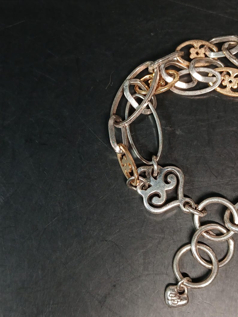 Puede incluir: Una pulsera plateada y dorada con una variedad de estilos de eslabones. La pulsera presenta eslabones ovalados, circulares y decorativos, incluido un cierre en forma de coraz&oacute;n. El metal tiene un acabado pulido.