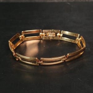 May include: A gold-colored bracelet with a rectangular link design. The bracelet is made of metal and has a clasp closure. The links are connected by small hinges. The bracelet is displayed on a dark surface.