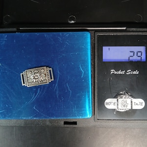 May include: A small, rectangular silver-toned pin with the words "FLOWERS HONOR CLUB" and a flower design. The pin is on a blue surface, next to a pocket scale displaying 2.9 grams.