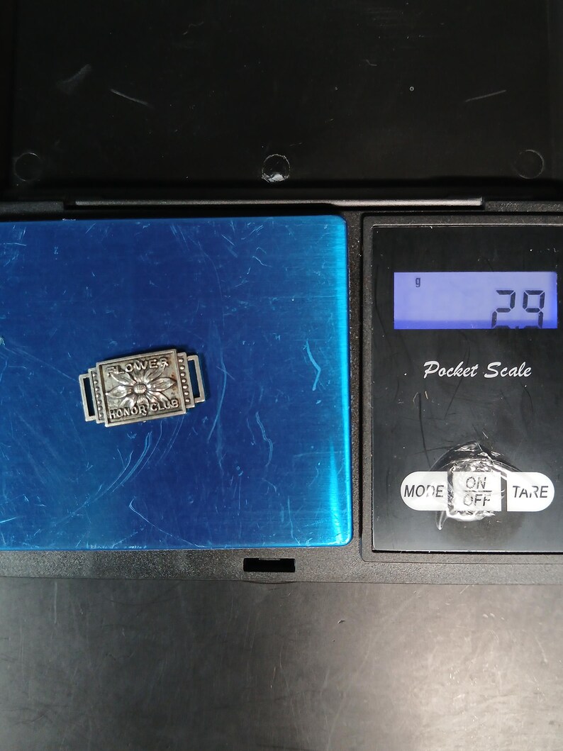 May include: A small, rectangular silver-toned pin with the words "FLOWERS HONOR CLUB" and a flower design. The pin is on a blue surface, next to a pocket scale displaying 2.9 grams.
