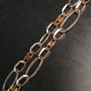 Puede incluir: Primer plano de un collar de cadena plateado y dorado. La cadena presenta eslabones ovalados alargados en plata, intercalados con eslabones decorativos dorados. El collar se muestra sobre un fondo oscuro, resaltando el contraste de los metales.