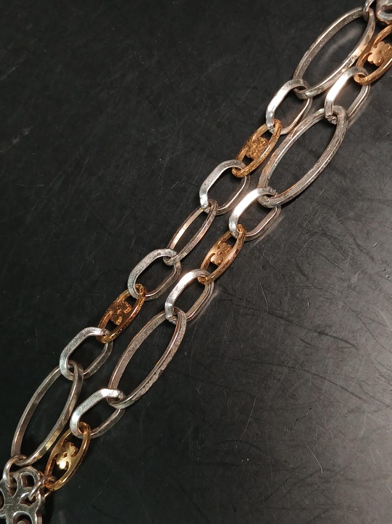 Puede incluir: Primer plano de un collar de cadena plateado y dorado. La cadena presenta eslabones ovalados alargados en plata, intercalados con eslabones decorativos dorados. El collar se muestra sobre un fondo oscuro, resaltando el contraste de los metales.