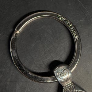 May include: Close-up of a silver-toned keychain with a large ring and decorative heart-shaped accents. The word "Brighton" is engraved on the ring. The keychain has an ornate design and is set against a dark background.