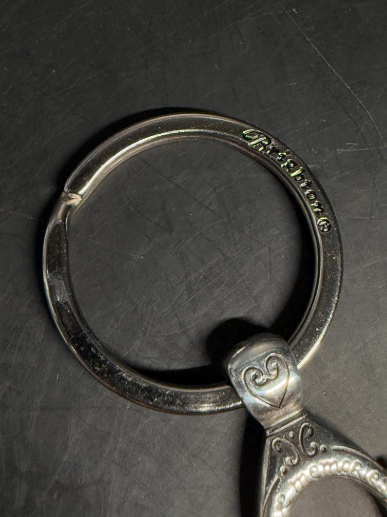 May include: Close-up of a silver-toned keychain with a large ring and decorative heart-shaped accents. The word "Brighton" is engraved on the ring. The keychain has an ornate design and is set against a dark background.
