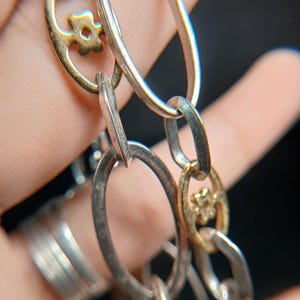 Puede incluir: Primer plano de una cadena plateada con eslabones ovalados y detalles dorados en forma de flor. La cadena est&aacute; compuesta por eslabones plateados y dorados alternados, creando un contraste de color y textura. El fondo es oscuro.