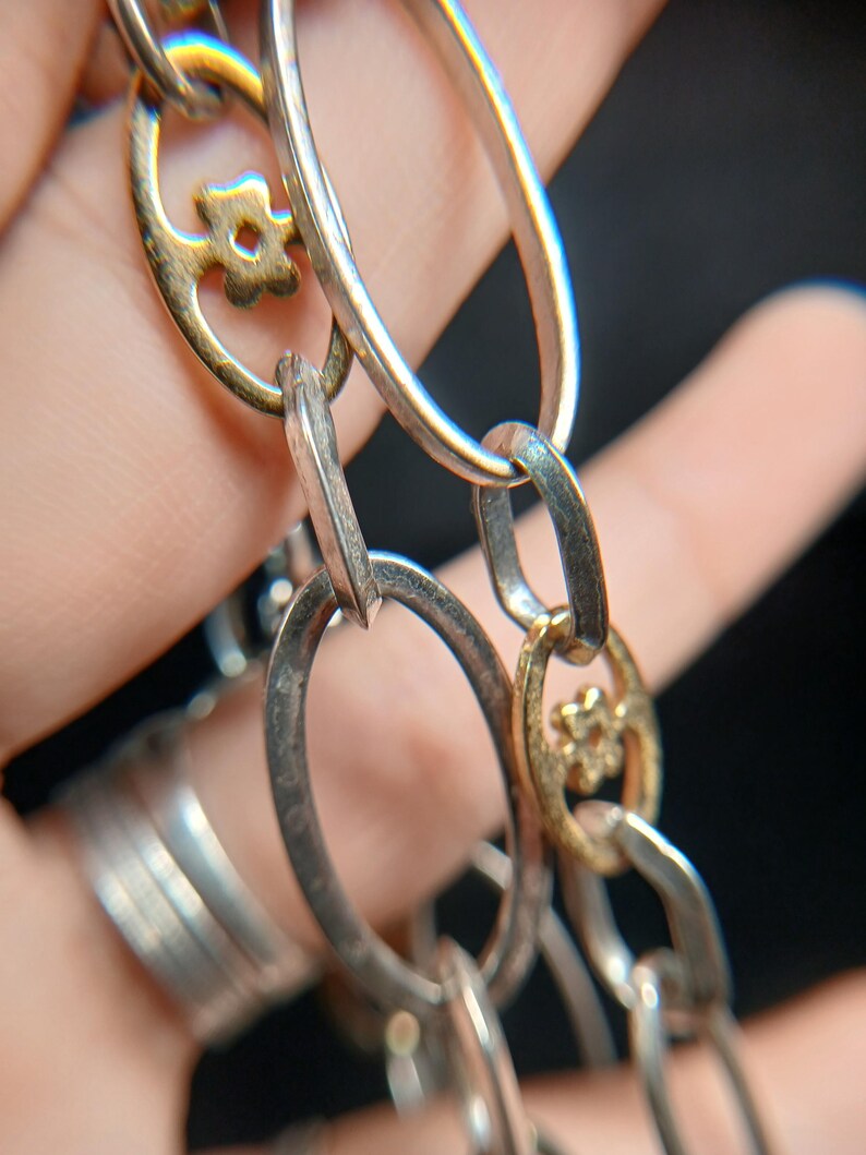 Puede incluir: Primer plano de una cadena plateada con eslabones ovalados y detalles dorados en forma de flor. La cadena est&aacute; compuesta por eslabones plateados y dorados alternados, creando un contraste de color y textura. El fondo es oscuro.