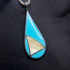 May include: A teardrop-shaped pendant featuring a bright turquoise inlay and a central triangular section with a shimmering, light-colored iridescent finish. The pendant is framed in silver and hangs from a matching silver chain link necklace.