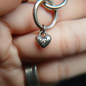 Puede incluir: Primer plano de una cadena de collar plateada con un dije en forma de coraz&oacute;n. El dije tiene una peque&ntilde;a letra "B" grabada. Los eslabones de la cadena son circulares y est&aacute;n conectados. El fondo est&aacute; borroso.