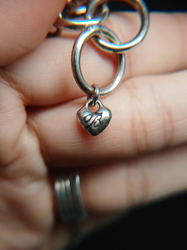 Puede incluir: Primer plano de una cadena de collar plateada con un dije en forma de coraz&oacute;n. El dije tiene una peque&ntilde;a letra "B" grabada. Los eslabones de la cadena son circulares y est&aacute;n conectados. El fondo est&aacute; borroso.