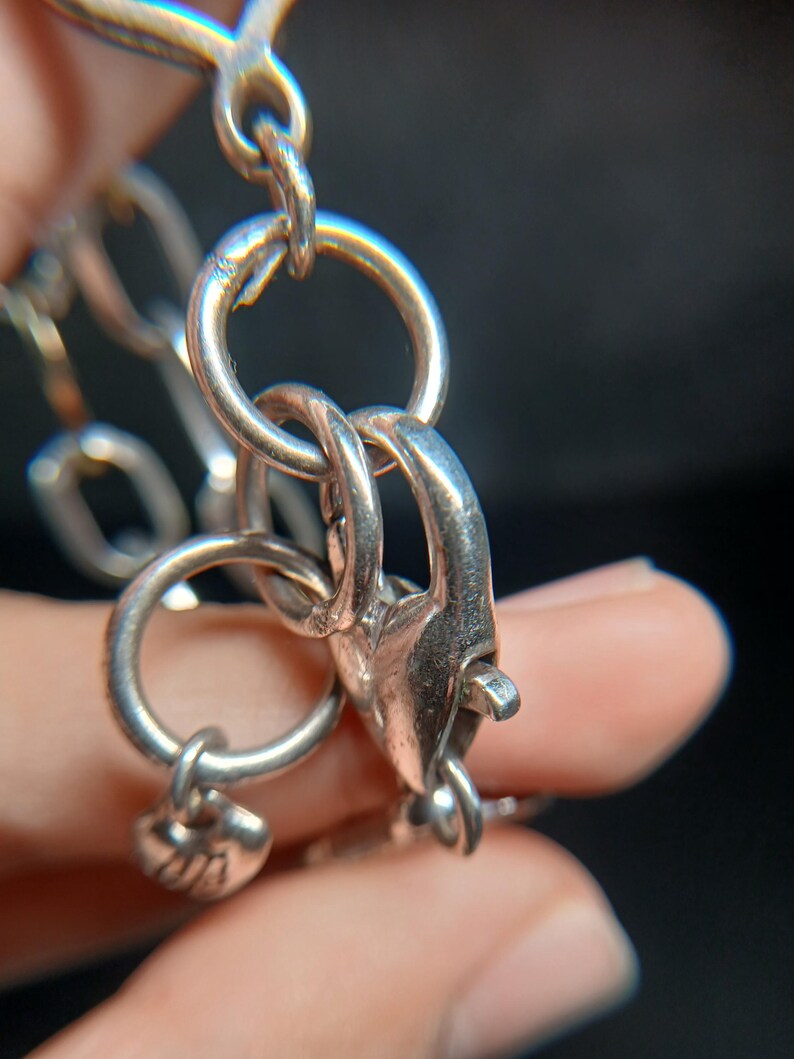 Puede incluir: Primer plano de un cierre de collar plateado y eslabones de cadena. El cierre es de estilo pinza de langosta, y los eslabones de la cadena son redondos. Un peque&ntilde;o dije est&aacute; unido a la cadena. La joyer&iacute;a es de metal brillante.