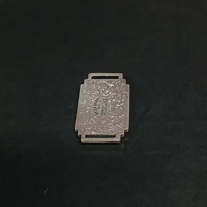 May include: A rectangular, silver-toned metal pendant with a textured surface. The pendant has a decorative, stepped design and a small loop at the top. The text "STERLING" is visible on the pendant.