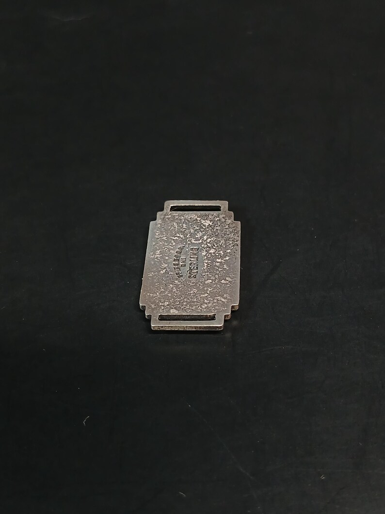 May include: A rectangular, silver-toned metal pendant with a textured surface. The pendant has a decorative, stepped design and a small loop at the top. The text "STERLING" is visible on the pendant.