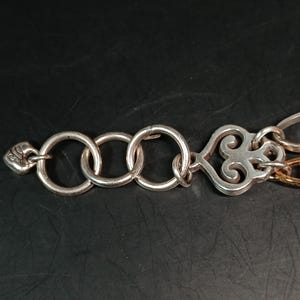 Puede incluir: Cadena de metal plateado con tres anillos entrelazados, un elemento decorativo en forma de coraz&oacute;n y un peque&ntilde;o cierre ornamentado. La cadena est&aacute; sobre una superficie oscura.