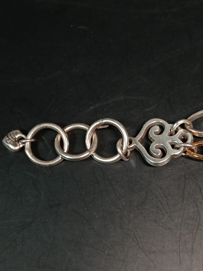 Puede incluir: Cadena de metal plateado con tres anillos entrelazados, un elemento decorativo en forma de coraz&oacute;n y un peque&ntilde;o cierre ornamentado. La cadena est&aacute; sobre una superficie oscura.