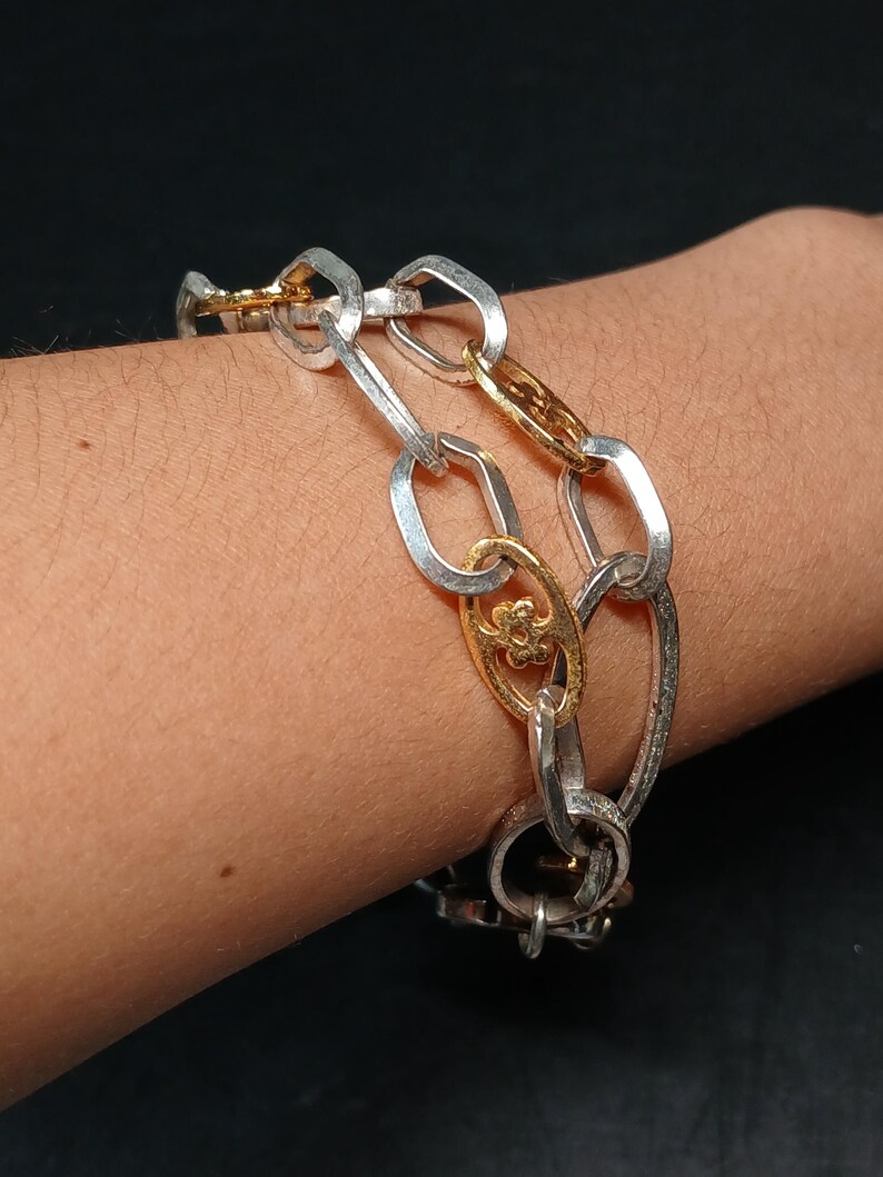 Puede incluir: Pulsera de cadena plateada y dorada. La pulsera presenta una mezcla de eslabones ovalados plateados y eslabones decorativos dorados. Los eslabones dorados tienen un dise&ntilde;o floral. La pulsera se lleva en la mu&ntilde;eca.
