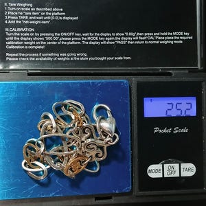 Puede incluir: Un collar plateado y dorado con un cierre en forma de coraz&oacute;n se muestra en una b&aacute;scula digital de bolsillo. La b&aacute;scula muestra un peso de 25,2 gramos. La b&aacute;scula tiene un bot&oacute;n de encendido/apagado y un bot&oacute;n de tara.