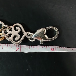 Puede incluir: Cadena de metal plateado con un cierre decorativo. El cierre tiene forma de coraz&oacute;n y est&aacute; conectado a una cadena con un dise&ntilde;o calado y ornamentado. Una cinta m&eacute;trica muestra que la cadena mide aproximadamente 18 cm de largo.