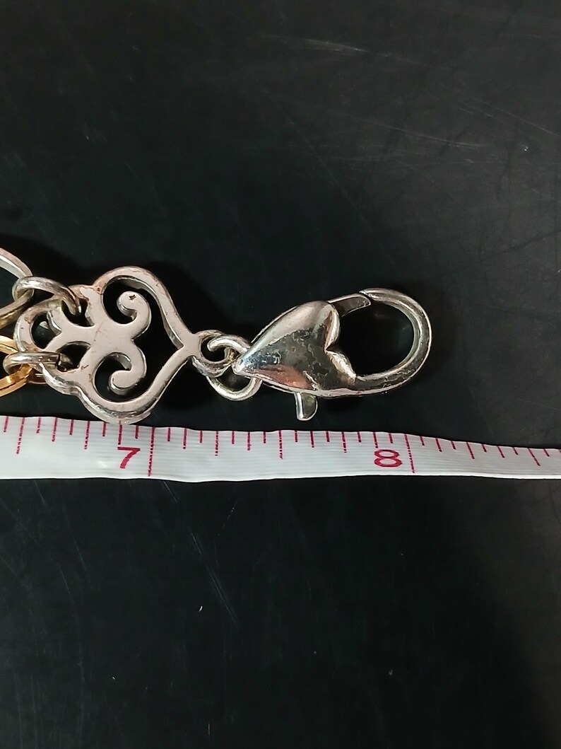 Puede incluir: Cadena de metal plateado con un cierre decorativo. El cierre tiene forma de coraz&oacute;n y est&aacute; conectado a una cadena con un dise&ntilde;o calado y ornamentado. Una cinta m&eacute;trica muestra que la cadena mide aproximadamente 18 cm de largo.