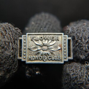 May include: A silver-toned metal pin with the words "FLOWER HONOR CLUB" and a flower design. The pin is rectangular with rounded edges and small circular details along the sides. The background is dark and out of focus.