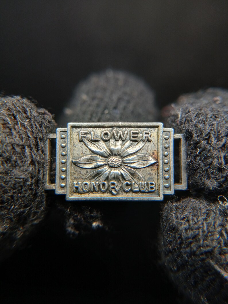 May include: A silver-toned metal pin with the words "FLOWER HONOR CLUB" and a flower design. The pin is rectangular with rounded edges and small circular details along the sides. The background is dark and out of focus.