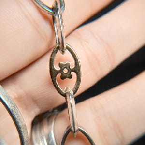 Puede incluir: Primer plano de una cadena plateada con un eslab&oacute;n ovalado dorado con un dise&ntilde;o floral. Los eslabones de la cadena son alargados y de forma ovalada. El fondo est&aacute; borroso, con un enfoque en la joyer&iacute;a.