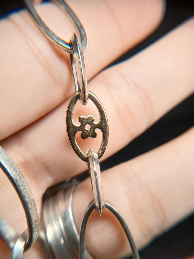 Puede incluir: Primer plano de una cadena plateada con un eslab&oacute;n ovalado dorado con un dise&ntilde;o floral. Los eslabones de la cadena son alargados y de forma ovalada. El fondo est&aacute; borroso, con un enfoque en la joyer&iacute;a.