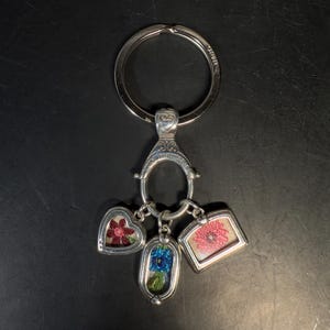 May include: Silver-toned keychain with three charms. The charms are a heart, an oval, and a rectangle, each containing a colorful floral design. The keychain has a large ring and a decorative clasp.