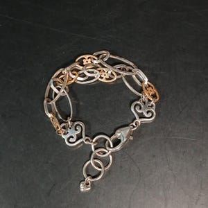 Puede incluir: Pulsera plateada con detalles dorados. La pulsera presenta una mezcla de eslabones ovalados y decorativos, incluyendo elementos en forma de coraz&oacute;n y ornamentados. Un cierre de mosquet&oacute;n asegura la pulsera.