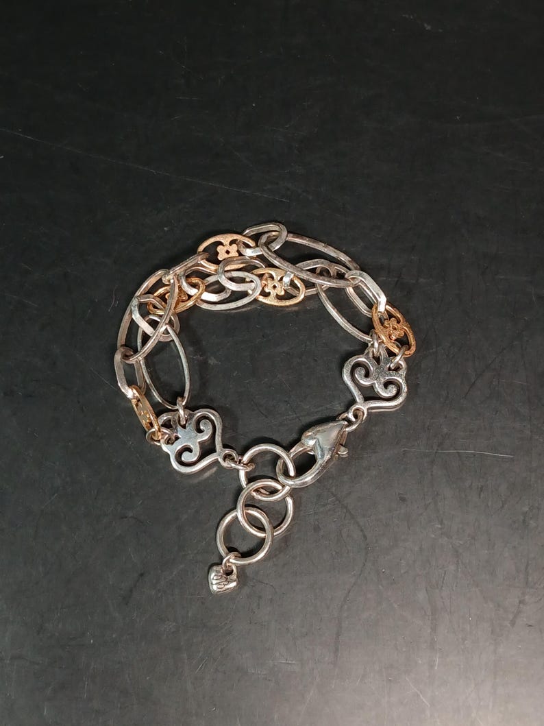 Puede incluir: Pulsera plateada con detalles dorados. La pulsera presenta una mezcla de eslabones ovalados y decorativos, incluyendo elementos en forma de coraz&oacute;n y ornamentados. Un cierre de mosquet&oacute;n asegura la pulsera.