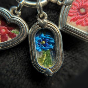 May include: Close-up of a silver charm bracelet with three detailed pendants. The pendants feature embroidered floral designs in shades of blue, pink, and red, set within ornate silver frames. The central pendant showcases a blue flower.