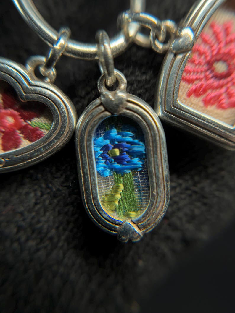 May include: Close-up of a silver charm bracelet with three detailed pendants. The pendants feature embroidered floral designs in shades of blue, pink, and red, set within ornate silver frames. The central pendant showcases a blue flower.