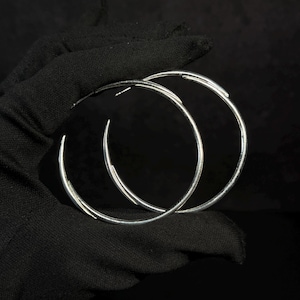 May include: A pair of large, polished silver hoop earrings with a thin, circular wire design. The earrings feature a smooth, reflective metallic finish and a simple, continuous loop structure that creates a clean, circular silhouette for the ears.