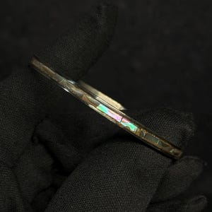 May include: A thin, circular bangle bracelet featuring a dark metal band inlaid with small, rectangular segments of iridescent abalone shell. The shell pieces display shifting hues of green, pink, and blue, creating a reflective, multi-colored pattern along the surface of the jewelry piece.